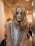 A mannequin with long blonde hair and dark eye makeup is dressed in a sleeveless white top featuring colorful patterns. It is positioned in a clothing store with a softly lit, modern interior.