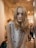 A mannequin with long blonde hair and dark eye makeup is dressed in a sleeveless white top featuring colorful patterns. It is positioned in a clothing store with a softly lit, modern interior.
