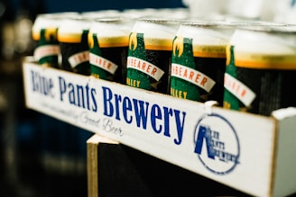 A close-up view of a cardboard carrier holding multiple beer cans with visible condensation. The carrier displays the brand name 'Blue Pants Brewery' in bold blue letters. The design on the cans is primarily green with red and white text.