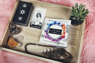 A wooden tray containing a variety of items arranged neatly. There is a deck of tarot cards with a card depicting a woman and a lion. The word 'Lumina Tarot' is printed on a visually appealing card with colorful watercolor splashes. Other items include a small Buddha statue, a crystal, a beaded bracelet, a feather, a circular object with the word 'YES', and a potted succulent plant. The tray is placed on a pink fur-like surface.