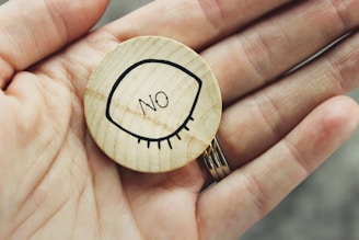 A person holds a small round wooden token. The token features a simple black line drawing of a closed eye with the word 'NO' inside it.
