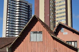 An old wooden house with a brown tiled roof stands in front of two modern high-rise apartment buildings. The house has a weathered appearance with a small window, and there is a 'for sale' sign on the side. The modern buildings behind are much taller, with multiple balconies and windows. The sky is a clear blue.