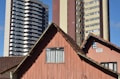 An old wooden house with a brown tiled roof stands in front of two modern high-rise apartment buildings. The house has a weathered appearance with a small window, and there is a 'for sale' sign on the side. The modern buildings behind are much taller, with multiple balconies and windows. The sky is a clear blue.