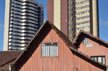 An old wooden house with a brown tiled roof stands in front of two modern high-rise apartment buildings. The house has a weathered appearance with a small window, and there is a 'for sale' sign on the side. The modern buildings behind are much taller, with multiple balconies and windows. The sky is a clear blue.