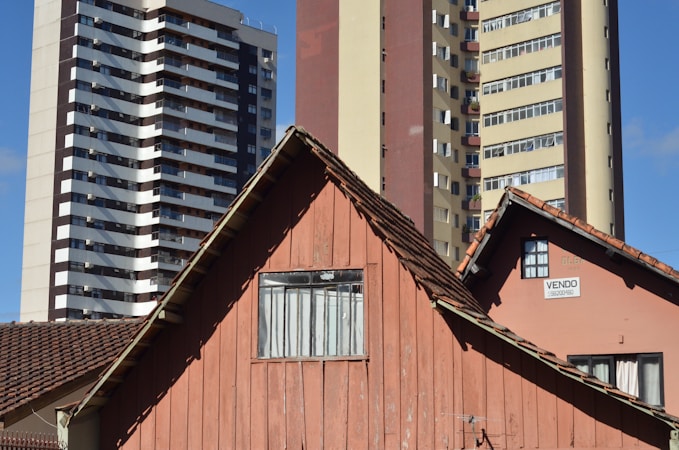 An old wooden house with a brown tiled roof stands in front of two modern high-rise apartment buildings. The house has a weathered appearance with a small window, and there is a 'for sale' sign on the side. The modern buildings behind are much taller, with multiple balconies and windows. The sky is a clear blue.