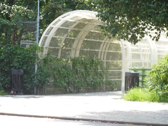 A white, mesh-covered arched structure surrounded by lush green foliage is visible. There are two black trash cans on each side near the entrance, and a green bench partially seen through the opening. A sign labeled Greenstreets is mounted on the fence.