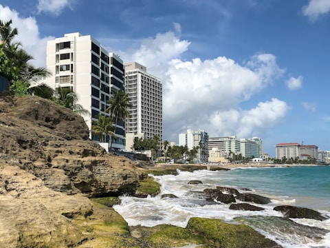 A coastal scene featuring modern high-rise buildings adjacent to a beach with rocks and palm trees. The sky is partly cloudy and the ocean waves are gently lapping against the shore.