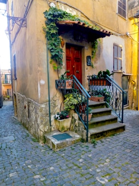 A charming, rustic building with stone walls and a red wooden door, complemented by a small staircase with wrought iron railings leading to the entrance. There are potted plants and flowers adorning the front, adding a touch of greenery to the scene. The cobblestone street enhances the historic and quaint ambiance.
