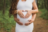 A pregnant woman dressed in a white form-fitting dress is in a forested area. Two hands, presumably hers, are placed on her belly, forming a heart shape with fingers. Another set of hands, likely from another person standing behind her, is placed below hers on the belly.