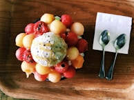 A wooden tray holds a dessert topped with a scoop of creamy ice cream and various colorful melon balls. Two spoons rest on a white napkin beside the dessert.