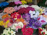 A vibrant array of various flower bouquets in a market setting, featuring a wide range of colors including red, pink, purple, yellow, and white. The flowers are arranged in clusters, creating a rich tapestry of textures and hues. Some bouquets are wrapped in clear plastic while others are placed directly in green buckets.