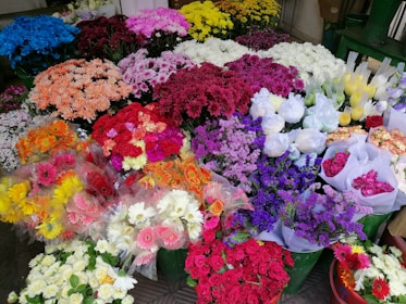 A vibrant array of various flower bouquets in a market setting, featuring a wide range of colors including red, pink, purple, yellow, and white. The flowers are arranged in clusters, creating a rich tapestry of textures and hues. Some bouquets are wrapped in clear plastic while others are placed directly in green buckets.