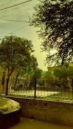 A serene urban scene featuring a fence with intricate design in front of a courtyard. Lush green trees partially block the sun, casting dappled light and shadows. A covered vehicle is parked on the left, and overhead cables cut across the sky.