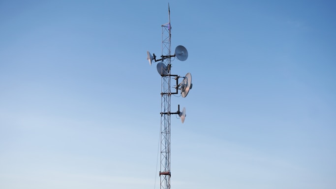 A tall communication tower is equipped with multiple satellite dishes and antennas against a clear blue sky.