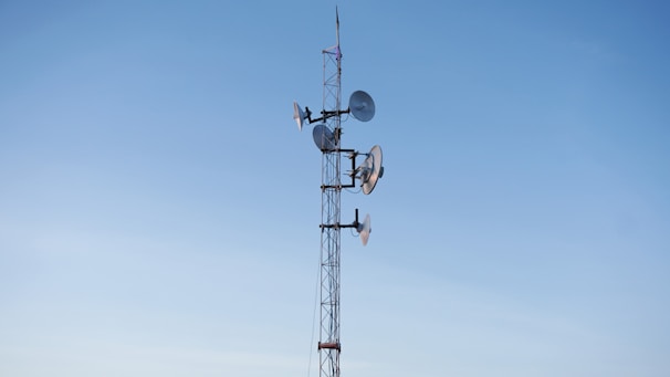 A tall communication tower is equipped with multiple satellite dishes and antennas against a clear blue sky.