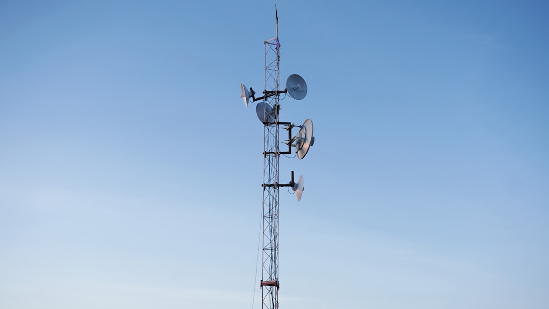 A tall communication tower is equipped with multiple satellite dishes and antennas against a clear blue sky.