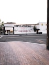 A street scene featuring a building with the sign 'TRIPLE A TRADING INC' prominently displayed. Additional signs offer services such as buying and selling gold, silver, guns, jewelry, and providing cash loans. The storefront has large windows and there's a brick sidewalk in the foreground leading to a quiet street. Few trees are visible around the area.