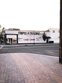 A street scene featuring a building with the sign 'TRIPLE A TRADING INC' prominently displayed. Additional signs offer services such as buying and selling gold, silver, guns, jewelry, and providing cash loans. The storefront has large windows and there's a brick sidewalk in the foreground leading to a quiet street. Few trees are visible around the area.