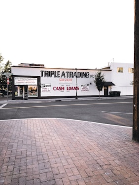 A street scene featuring a building with the sign 'TRIPLE A TRADING INC' prominently displayed. Additional signs offer services such as buying and selling gold, silver, guns, jewelry, and providing cash loans. The storefront has large windows and there's a brick sidewalk in the foreground leading to a quiet street. Few trees are visible around the area.