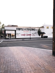 A street scene featuring a building with the sign 'TRIPLE A TRADING INC' prominently displayed. Additional signs offer services such as buying and selling gold, silver, guns, jewelry, and providing cash loans. The storefront has large windows and there's a brick sidewalk in the foreground leading to a quiet street. Few trees are visible around the area.