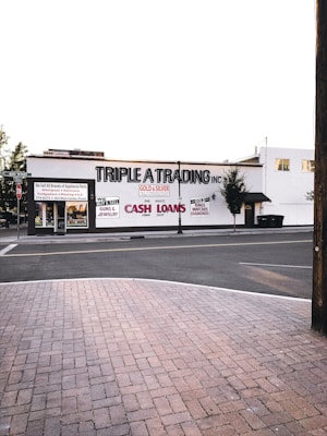 A street scene featuring a building with the sign 'TRIPLE A TRADING INC' prominently displayed. Additional signs offer services such as buying and selling gold, silver, guns, jewelry, and providing cash loans. The storefront has large windows and there's a brick sidewalk in the foreground leading to a quiet street. Few trees are visible around the area.