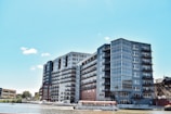Modern waterfront buildings with multiple stories, featuring large windows and balconies. A boat is docked along the waterway, with several trees and a clear blue sky in the background.