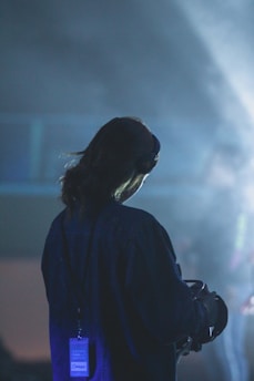 A person stands with their back facing the camera, wearing a lanyard and holding what appears to be a VR headset. The lighting is dim, with a bluish hue creating a mysterious and ethereal ambiance. There is a soft spotlight highlighting the person against a blurred background.