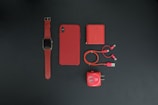 A collection of red tech accessories neatly arranged on a dark background. Items include a smartwatch with a red strap, a smartphone with a red case, a red power bank, red charging cables, and a red USB wall adapter. The minimalist composition emphasizes the vibrant red color theme.