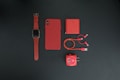 A collection of red tech accessories neatly arranged on a dark background. Items include a smartwatch with a red strap, a smartphone with a red case, a red power bank, red charging cables, and a red USB wall adapter. The minimalist composition emphasizes the vibrant red color theme.
