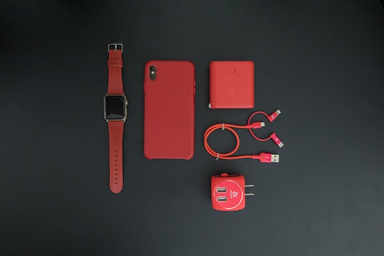A collection of red tech accessories neatly arranged on a dark background. Items include a smartwatch with a red strap, a smartphone with a red case, a red power bank, red charging cables, and a red USB wall adapter. The minimalist composition emphasizes the vibrant red color theme.