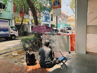A street artist sits on a small stool, drawing on a large canvas supported by an easel. He is in a shaded area on a sidewalk under a tree. Nearby are several colorful buildings with commercial signs in English and other languages. Vehicles are parked along the street, including a white van and a car. The scene includes a parked bicycle and a backpack on the ground.