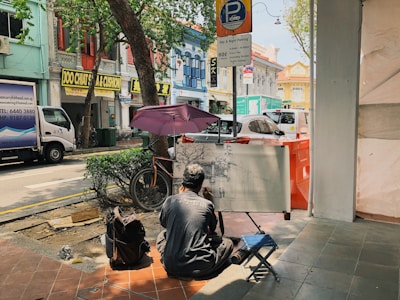 A street artist sits on a small stool, drawing on a large canvas supported by an easel. He is in a shaded area on a sidewalk under a tree. Nearby are several colorful buildings with commercial signs in English and other languages. Vehicles are parked along the street, including a white van and a car. The scene includes a parked bicycle and a backpack on the ground.