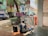 A street artist sits on a small stool, drawing on a large canvas supported by an easel. He is in a shaded area on a sidewalk under a tree. Nearby are several colorful buildings with commercial signs in English and other languages. Vehicles are parked along the street, including a white van and a car. The scene includes a parked bicycle and a backpack on the ground.