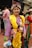 A person wearing traditional attire, featuring a vibrant combination of colors and textures. The outfit includes a yellow sash, an ornate headdress adorned with beads, and a purple robe with intricate patterns. The background shows a group of people gathered together outdoors, suggesting a social or cultural event.