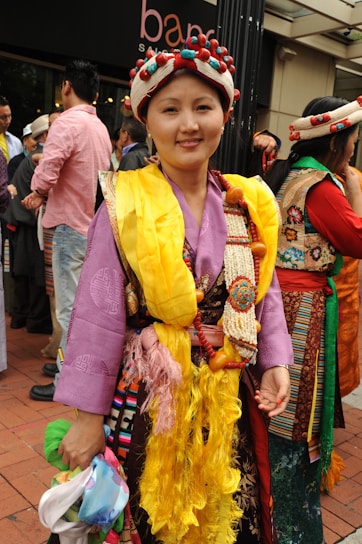 A person wearing traditional attire, featuring a vibrant combination of colors and textures. The outfit includes a yellow sash, an ornate headdress adorned with beads, and a purple robe with intricate patterns. The background shows a group of people gathered together outdoors, suggesting a social or cultural event.