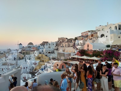 A picturesque view of a coastal town with whitewashed buildings and dome-shaped roofs, likely on a Greek island. The architecture overlooks the sea and is bathed in soft evening light. Numerous people are gathered, possibly tourists, enjoying the scenery and taking photos. A traditional windmill is visible, adding to the charm of the scene.