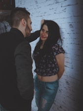 A couple stands close together against a white brick wall. The woman is smiling and leaning slightly against the wall with her hands behind her back, while the man faces her, appearing engaged in conversation. She wears a floral top and ripped jeans, and he is dressed in a dark jacket.