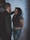 A couple stands close together against a white brick wall. The woman is smiling and leaning slightly against the wall with her hands behind her back, while the man faces her, appearing engaged in conversation. She wears a floral top and ripped jeans, and he is dressed in a dark jacket.