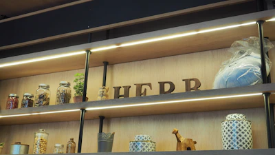 Wooden shelves hold various items, including jars of dried goods, a potted plant, and decorative objects. The word 'HERB' is spelled out with wooden letters, and there is a wrapped package. The layout is illuminated with accent lighting.