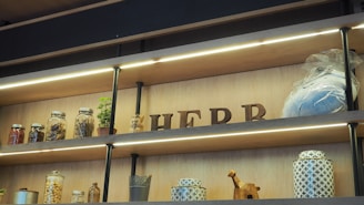 Wooden shelves hold various items, including jars of dried goods, a potted plant, and decorative objects. The word 'HERB' is spelled out with wooden letters, and there is a wrapped package. The layout is illuminated with accent lighting.