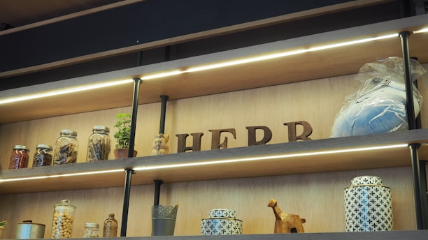Wooden shelves hold various items, including jars of dried goods, a potted plant, and decorative objects. The word 'HERB' is spelled out with wooden letters, and there is a wrapped package. The layout is illuminated with accent lighting.