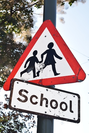 A triangular warning road sign with a red border and white background depicts two silhouetted figures, possibly children, holding hands. Below it, there's a rectangular sign with the word 'School' in bold black letters. The signs are mounted on a pole, situated amidst tree foliage with visible branches and leaves.