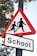 A triangular warning road sign with a red border and white background depicts two silhouetted figures, possibly children, holding hands. Below it, there's a rectangular sign with the word 'School' in bold black letters. The signs are mounted on a pole, situated amidst tree foliage with visible branches and leaves.