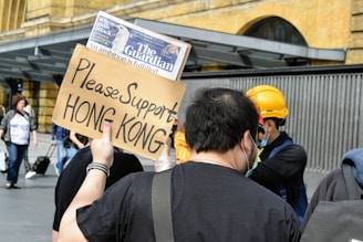 A group of people is gathered outdoors, with one person holding a cardboard sign that reads 'Please Support HONG KONG.' A person nearby is wearing a yellow construction helmet and a mask. The scene takes place near a building facade with visible pedestrians in the background.