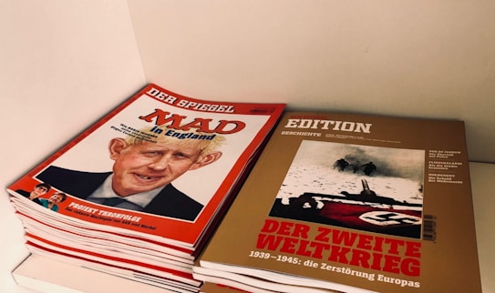 A stack of magazines is pictured, with the top left one featuring a caricature of a man with a headline in red and white, while the top right magazine has a historical image with prominent Nazi imagery and a headline about World War II.