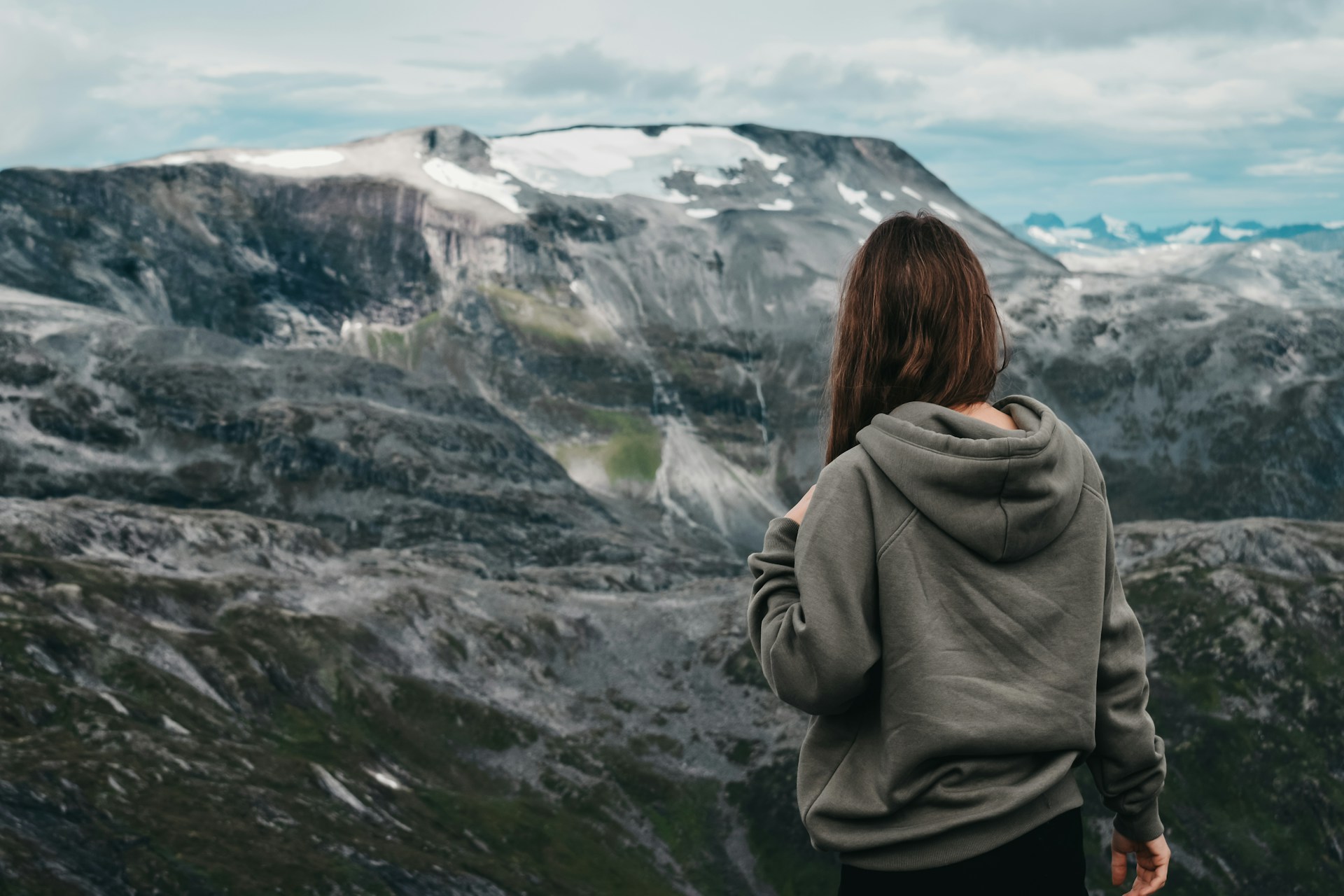 woman wearing gray hooded jacket standing and facing back on mountain under blue and white skies during daytime