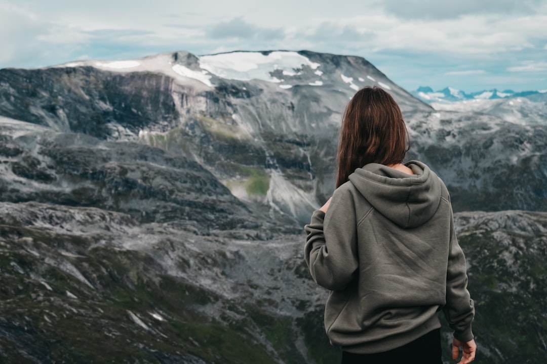 woman wearing gray hooded jacket standing and facing back on mountain under blue and white skies during daytime