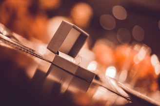 An elegant engagement ring sits in an open jewelry box, reflecting light from a softly blurred background. The setting creates a romantic and warm ambiance.