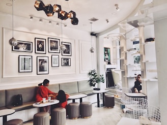 A modern cafe interior with white walls, featuring a series of black-and-white photos arranged on the wall. The space includes cozy seating with light-colored cushions and circular tables. Two people are seated in conversation on the left side, and another person is visible in the background near tall white shelves. The lighting is elegant with geometric fixtures and spherical lamps.