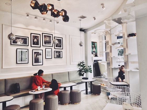 A modern cafe interior with white walls, featuring a series of black-and-white photos arranged on the wall. The space includes cozy seating with light-colored cushions and circular tables. Two people are seated in conversation on the left side, and another person is visible in the background near tall white shelves. The lighting is elegant with geometric fixtures and spherical lamps.